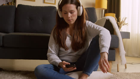 Woman-Wearing-Wireless-Headphones-Sitting-On-Floor-At-Home-Streaming-Music-From-Mobile-Phone-4